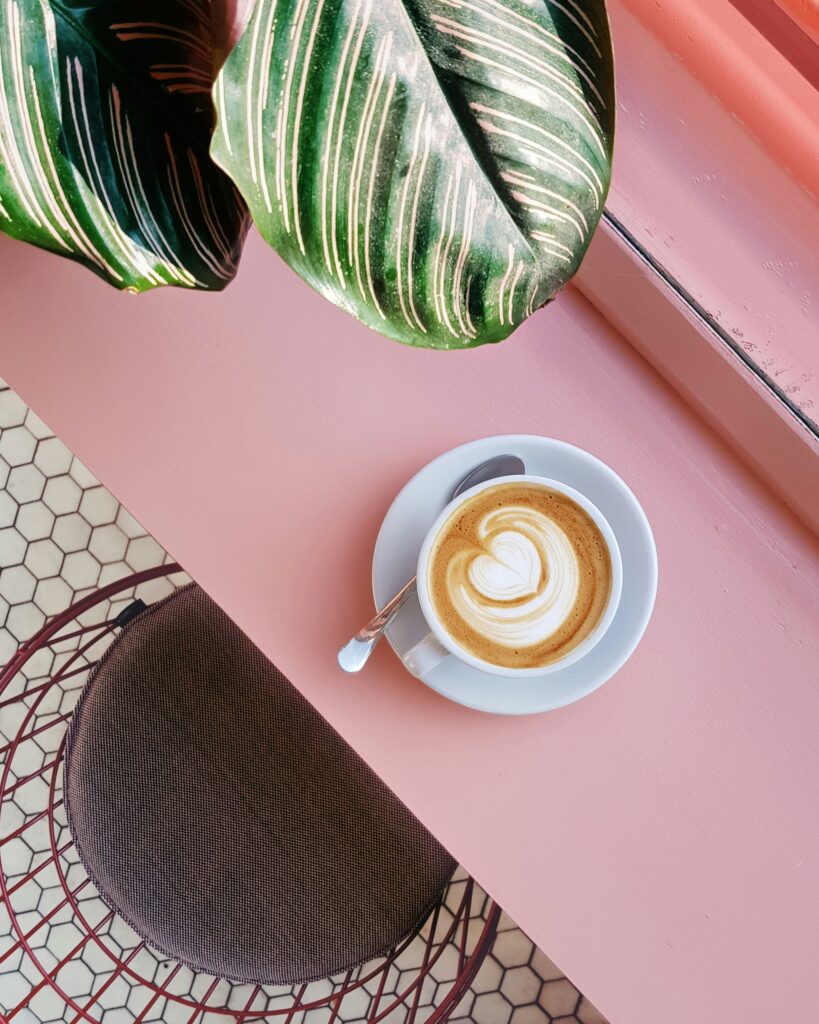 Latte mit Herz im Milchschaum auf rosa Tisch, daneben Löffel und grüne Blätter.