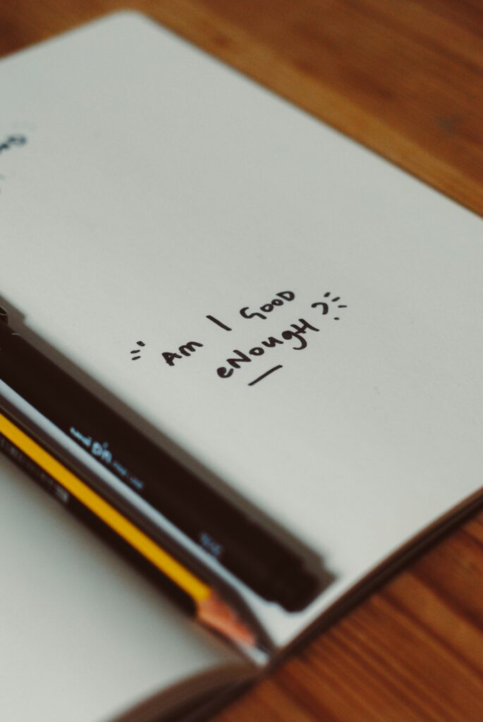 Notizbuch mit handgeschriebener Frage ‚Am I good enough?‘ sowie Stift und Bleistift.
